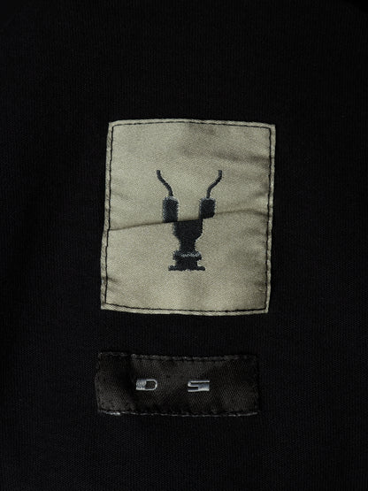 Close-up of a black garment featuring a rectangular beige patch with a black embroidered deer head logo and a black sewn-in size label below it.