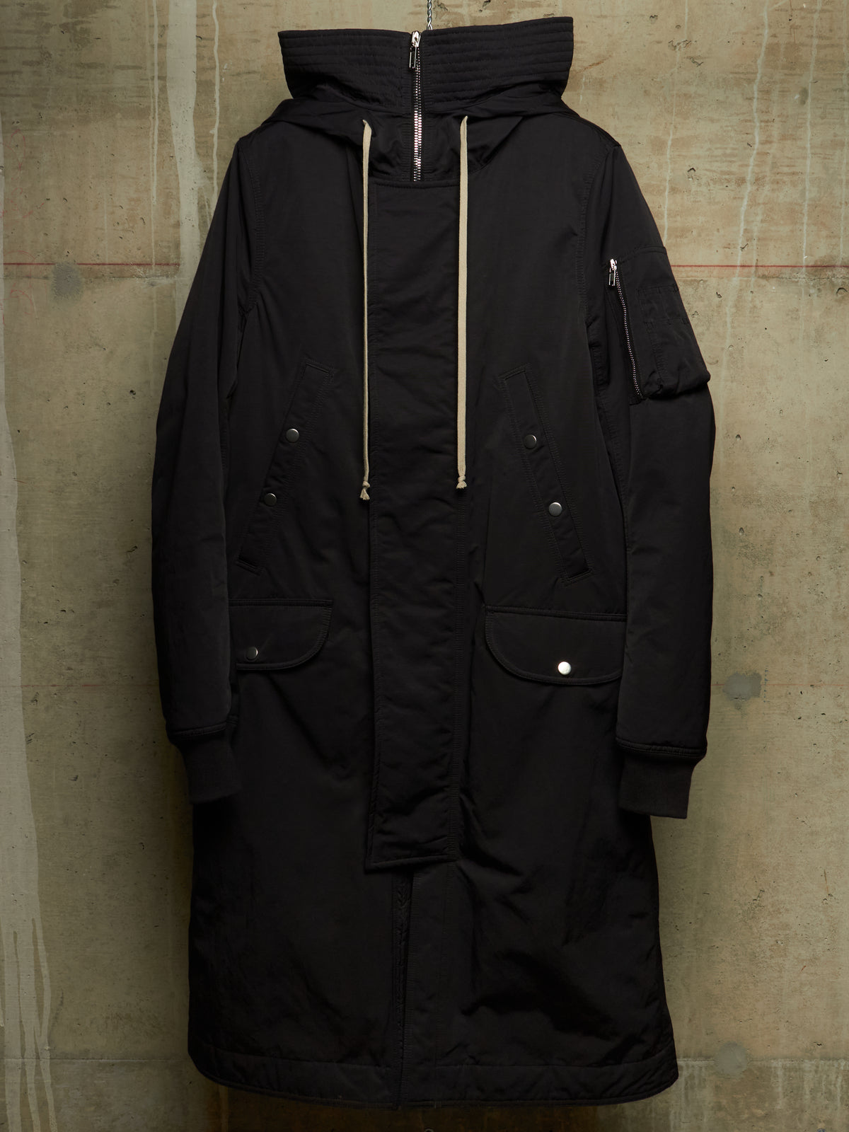 A long black hooded parka jacket with beige drawstrings, displayed hanging against a textured concrete wall.