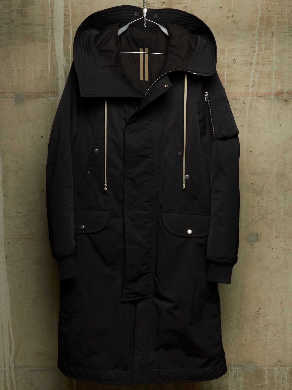 A black hooded parka jacket with gold-toned zipper details and silver snap buttons, hanging against a textured concrete wall.