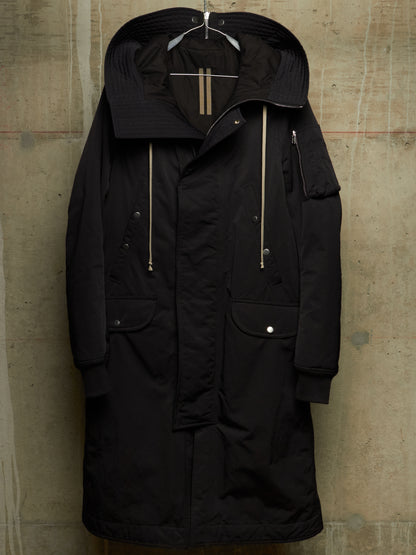 A black hooded parka jacket with gold-toned zipper details and silver snap buttons, hanging against a textured concrete wall.