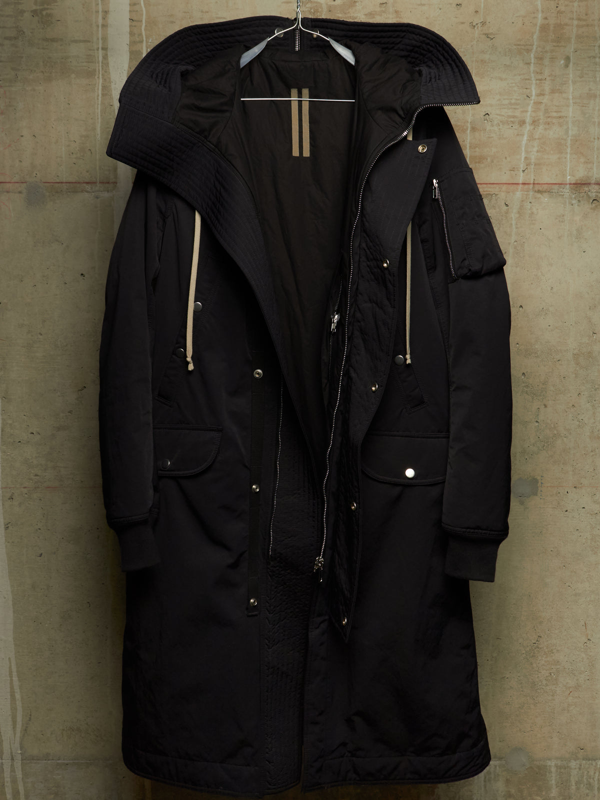 A black hooded parka coat hanging on a hanger against a textured concrete wall. The coat features a ribbed knit hood, front zipper and snap closure, zippered pockets, and light beige drawstrings.