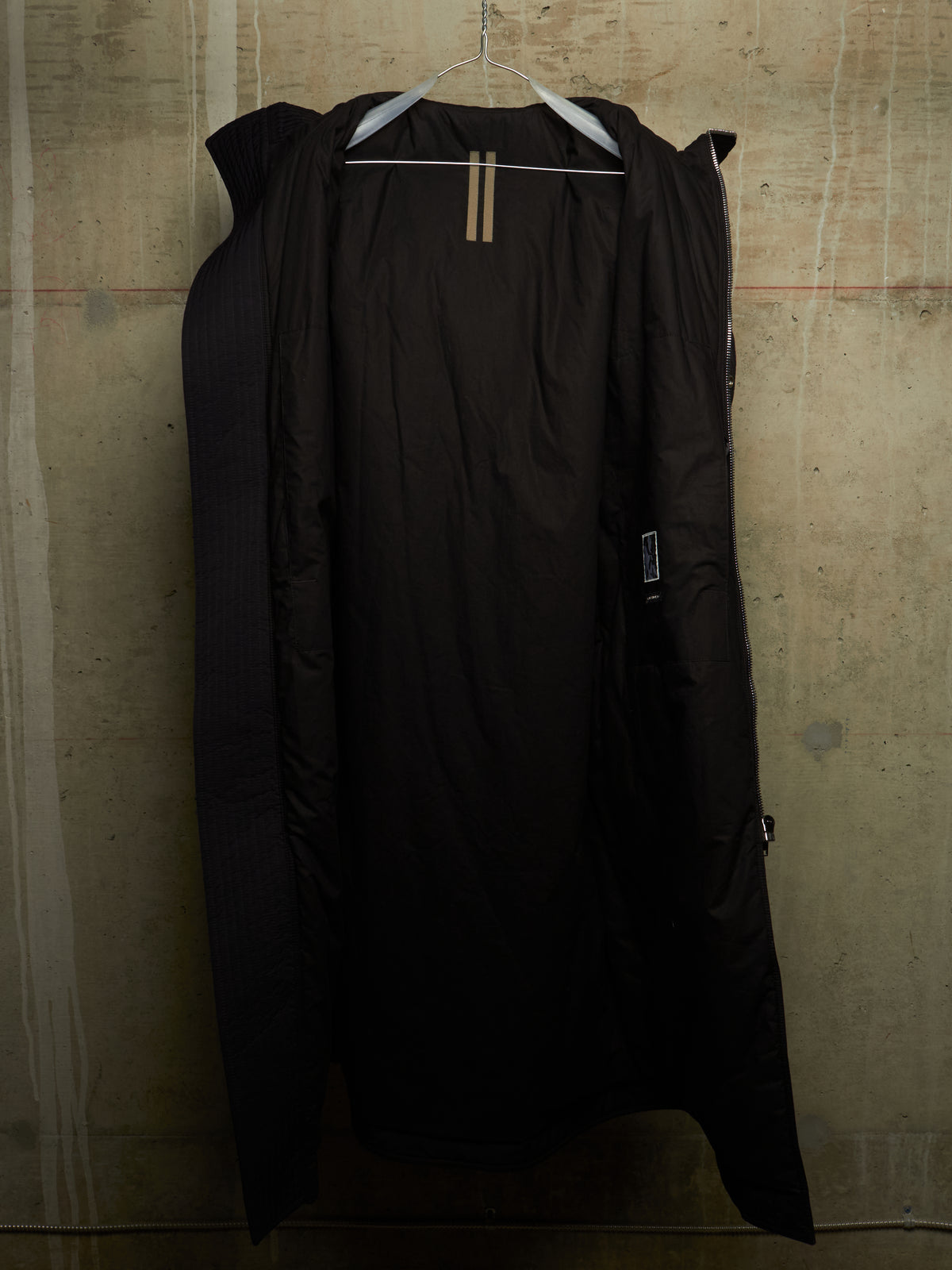 A long black puffer coat hanging on a white hanger against a textured concrete wall.