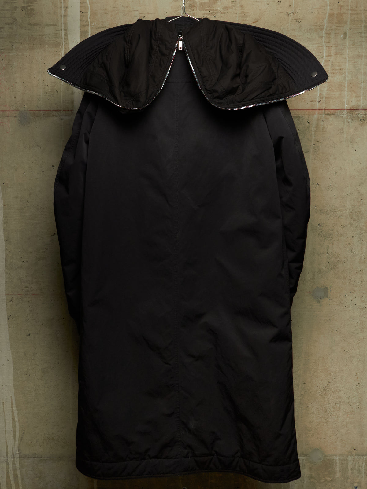 A black oversized coat with a wide, structured collar, hanging on a metal hanger against a textured concrete wall.
