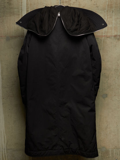 A black oversized coat with a wide, structured collar, hanging on a metal hanger against a textured concrete wall.