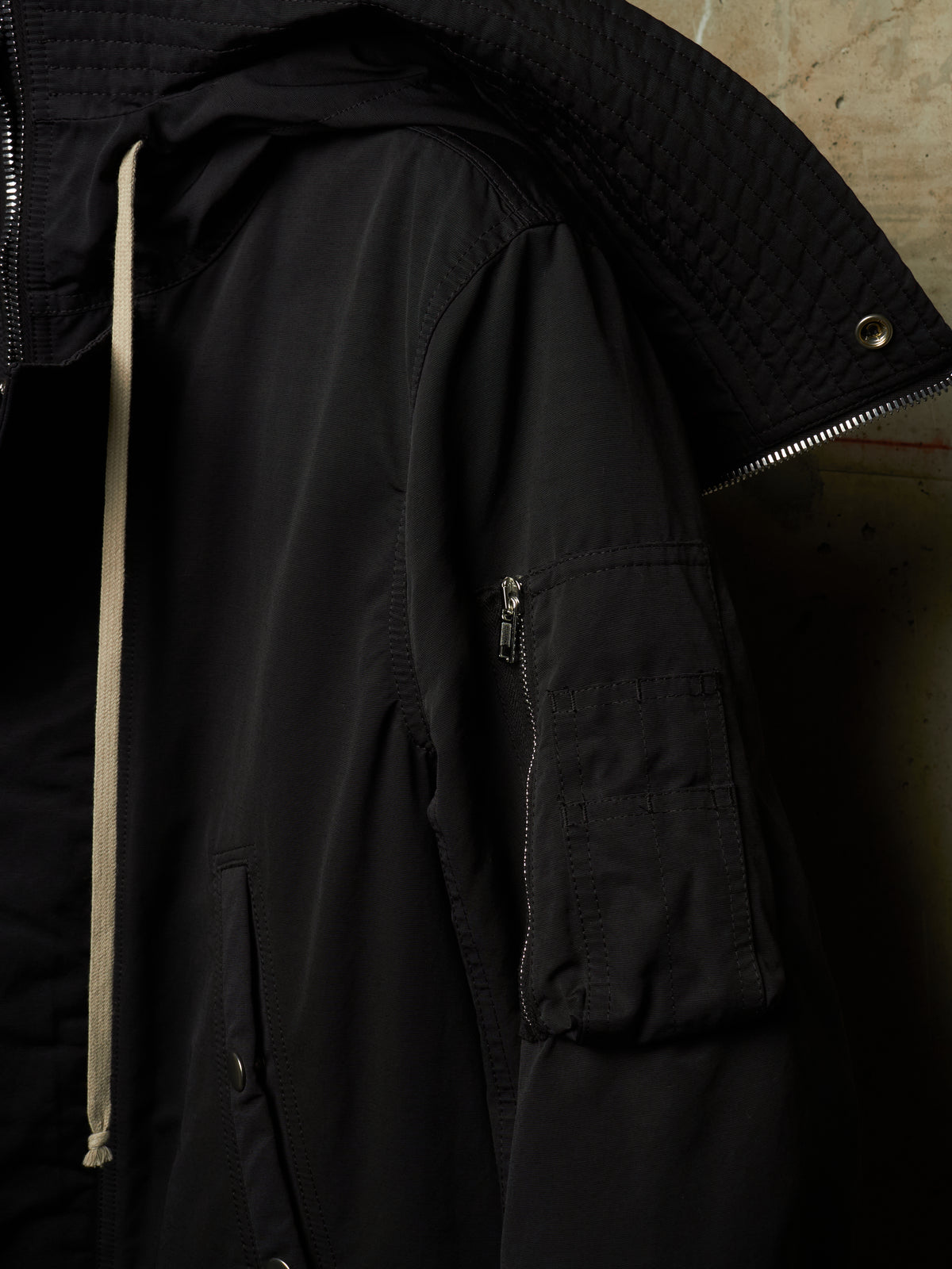 Close-up of a black hooded jacket with a quilted hood, a beige drawstring, and a zippered sleeve pocket, set against a dark textured background.