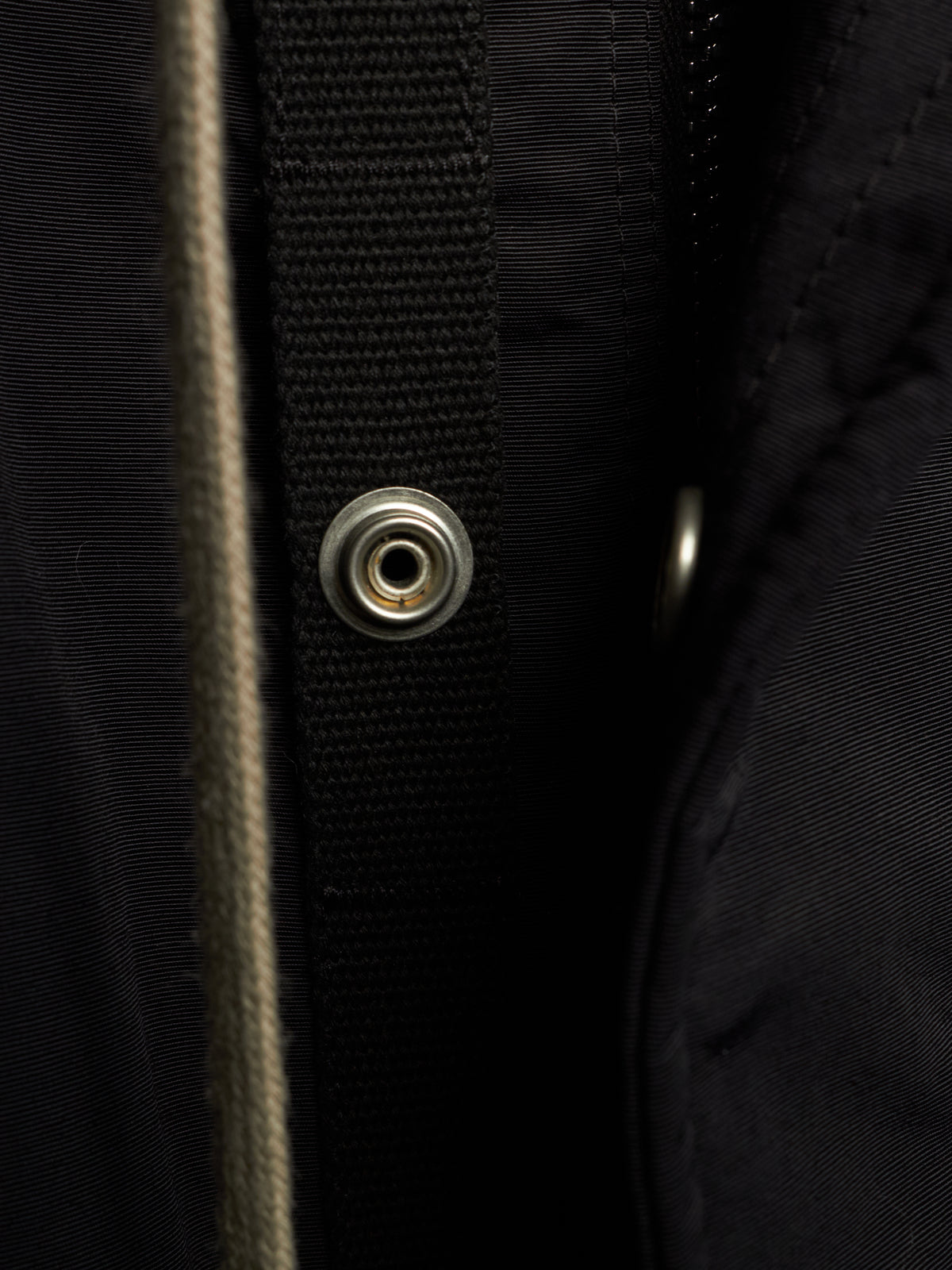 Close-up detail of a black jacket showing a black webbing strap with a silver metal snap button, a vertical beige zipper, and dark textured fabric.