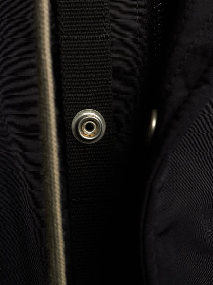 Close-up detail of a black jacket showing a black webbing strap with a silver metal snap button, a vertical beige zipper, and dark textured fabric.