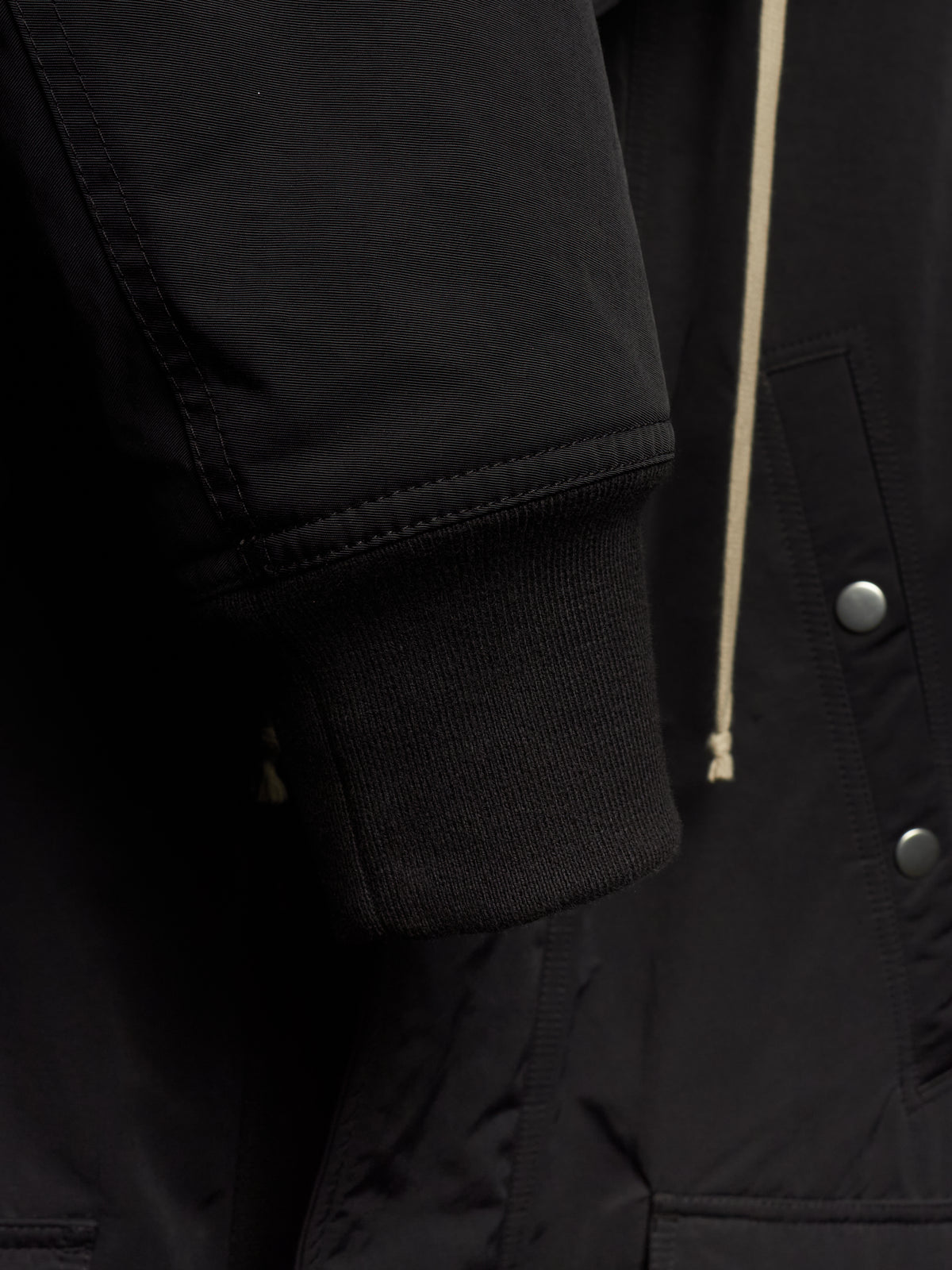 Close-up detail of a black jacket showing the ribbed knit cuff, a cream-colored drawstring, and silver snap buttons.