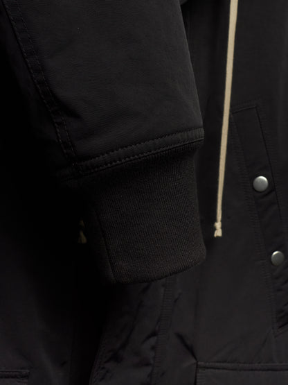Close-up detail of a black jacket showing the ribbed knit cuff, a cream-colored drawstring, and silver snap buttons.