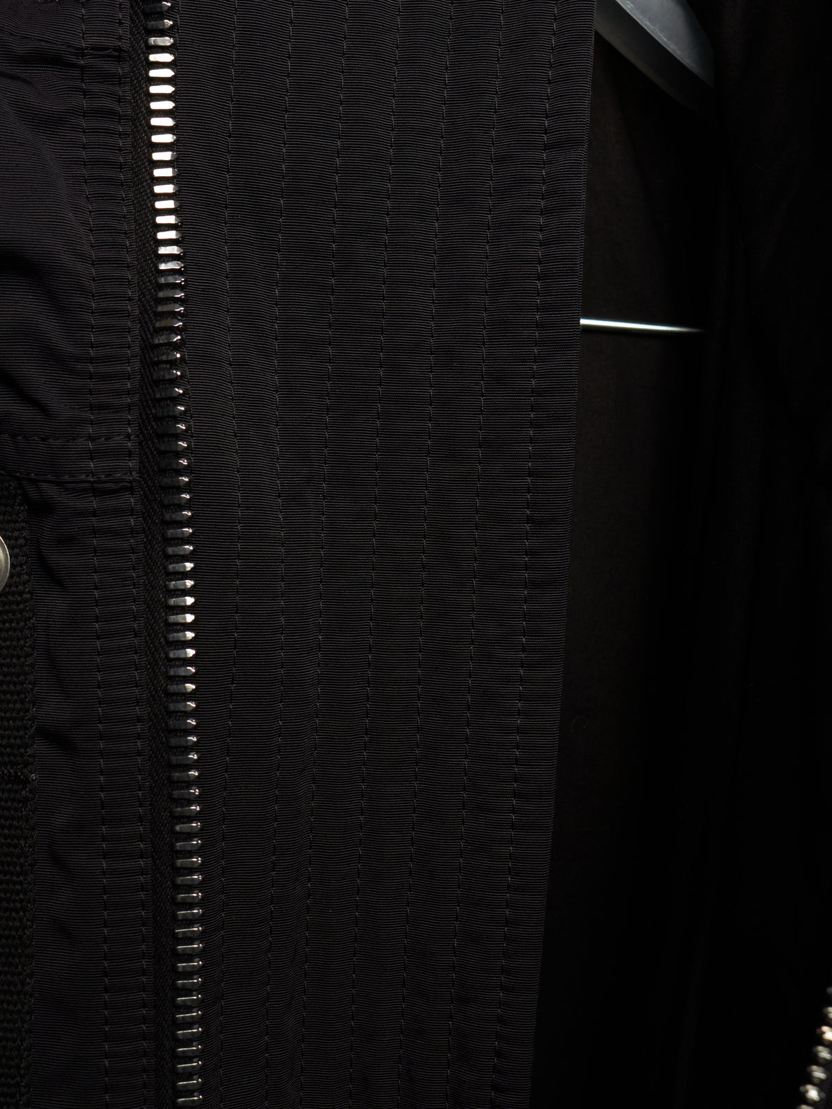 Close-up detail of a black jacket featuring a prominent silver zipper running vertically down the front, with textured stitching and a dark interior lining.