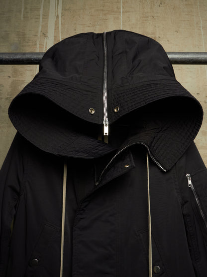 A black hooded jacket with a wide brimmed hood, quilted texture, and silver zippers, hanging against a textured concrete wall.
