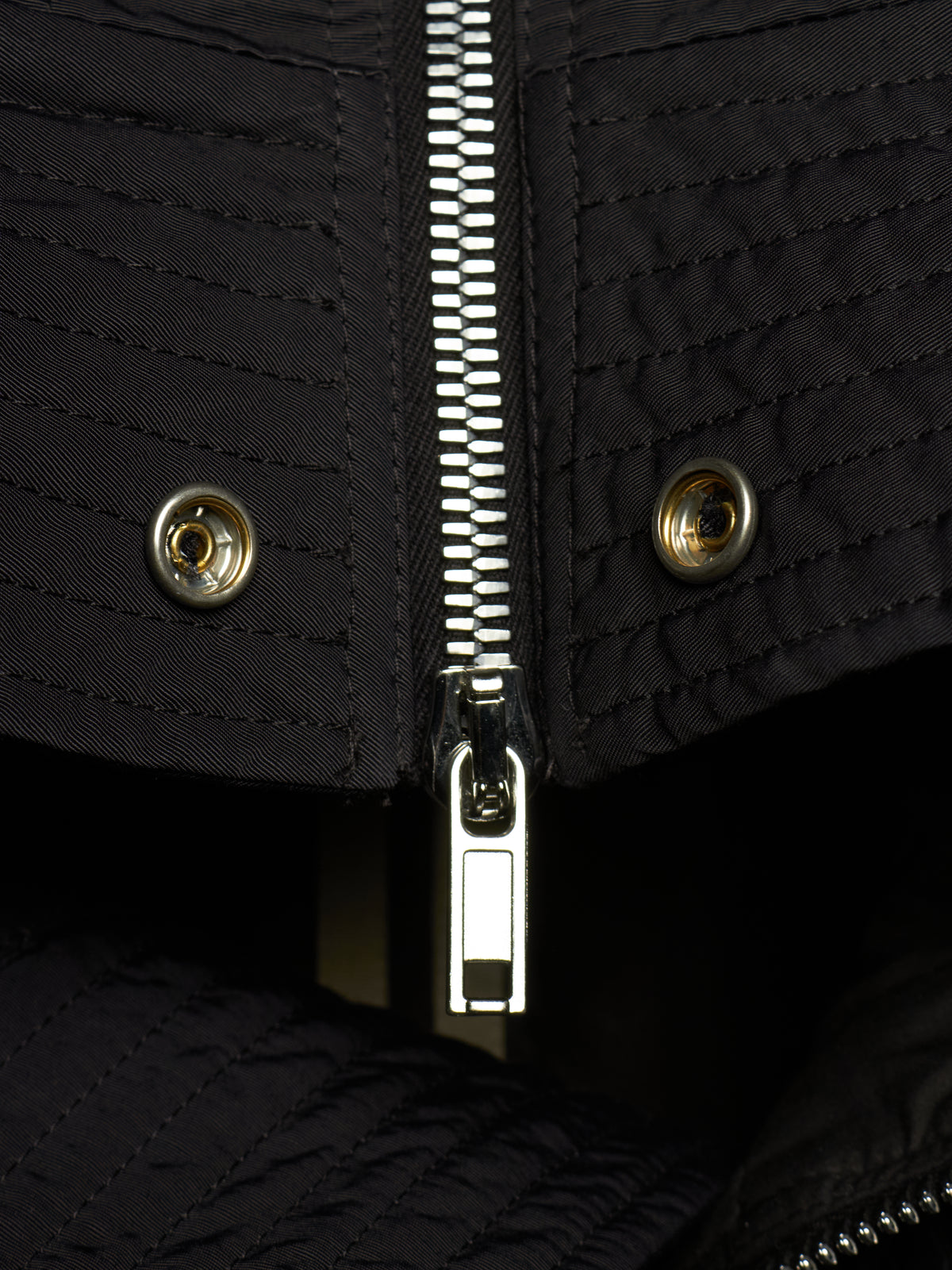 Close-up of a black quilted jacket featuring a central silver metal zipper, two matching snap buttons on the flap, and a dark interior lining.