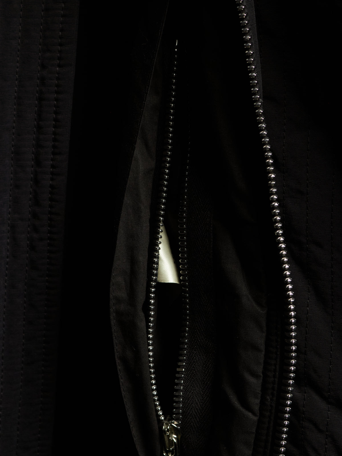 Close-up of a black quilted jacket with a silver metal beaded zipper and a small white triangular tab visible inside the pocket.