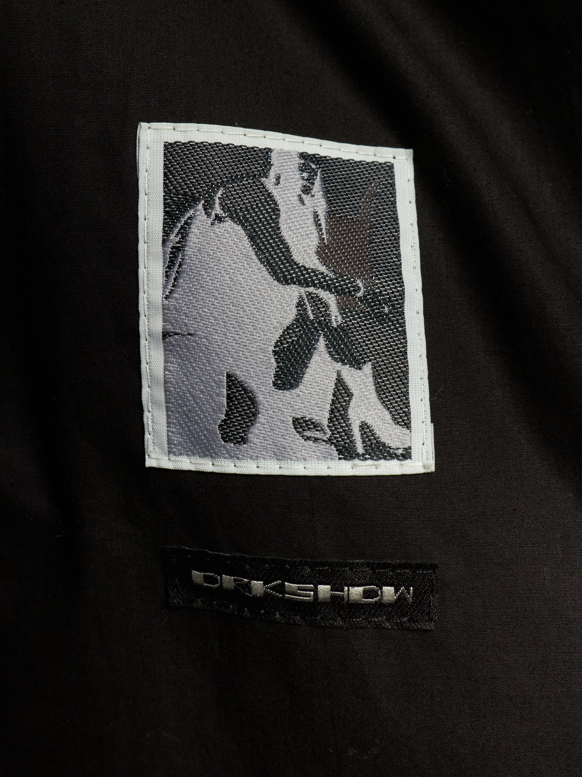 Close-up of a black garment featuring a rectangular embroidered patch with a grayscale horse design and a black brand label below reading ORKSHOW.