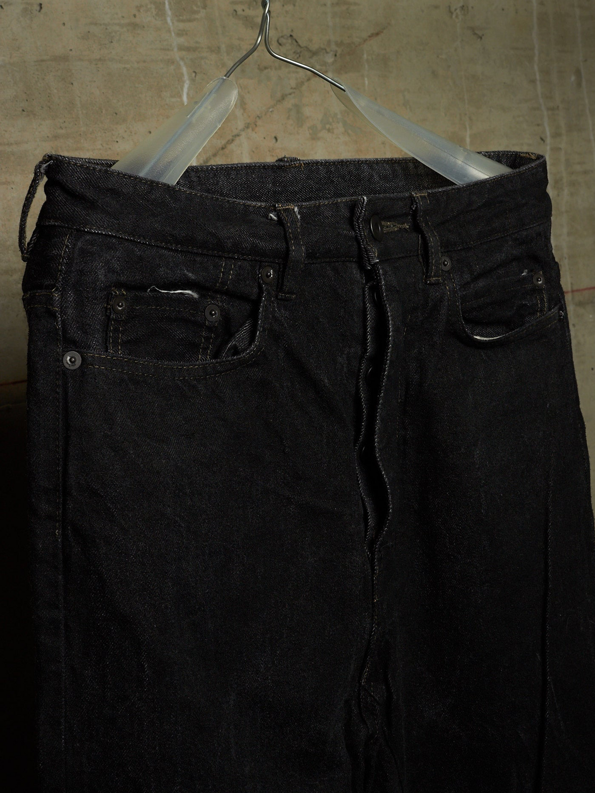 Black denim jeans hanging on a clear plastic hanger against a textured concrete wall.