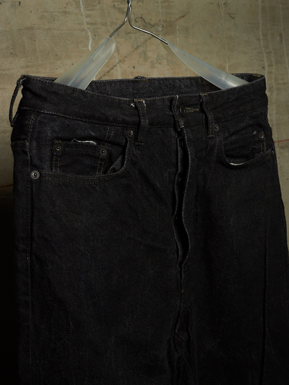 Black denim jeans hanging on a clear plastic hanger against a textured concrete wall.