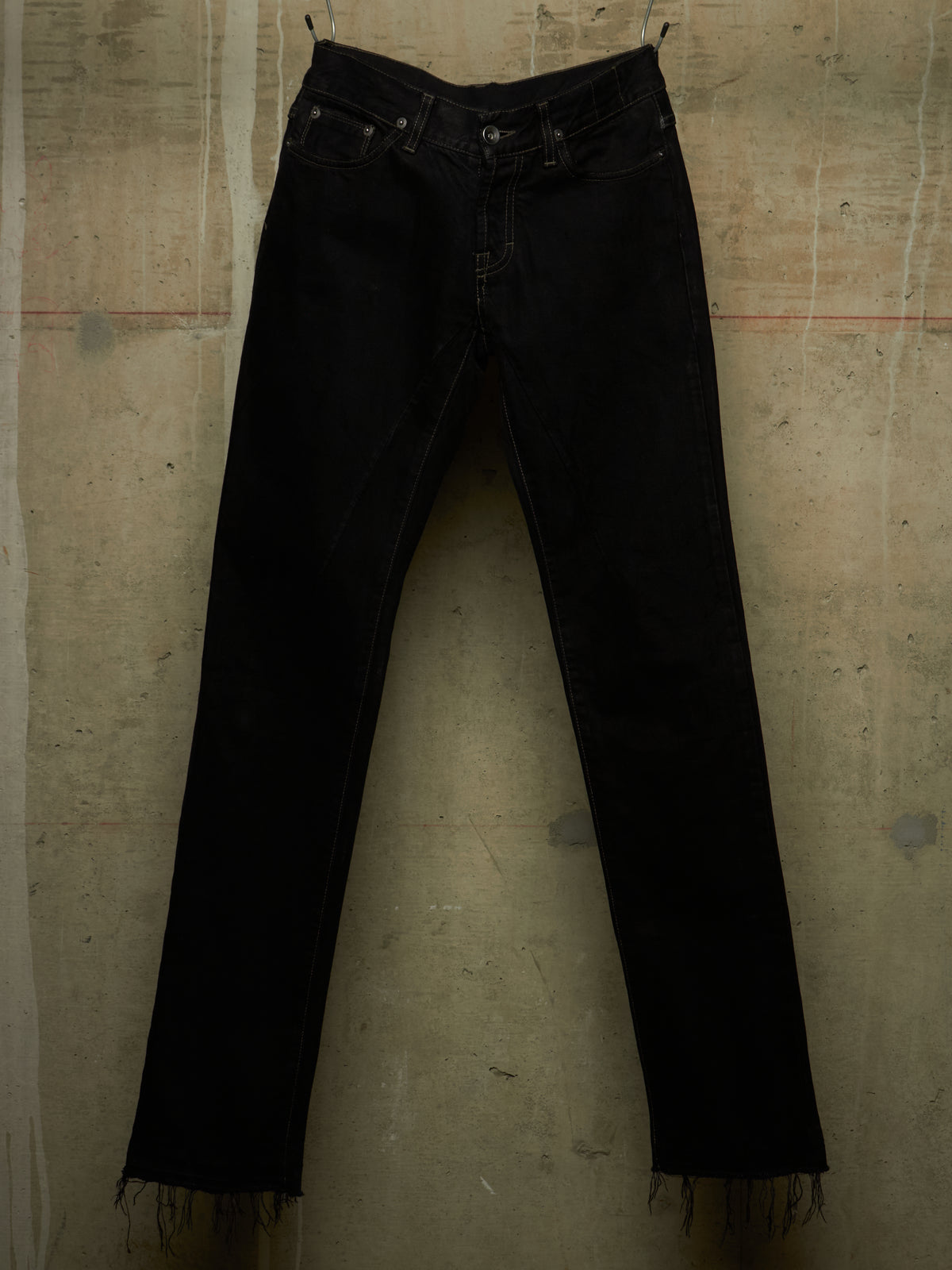 Black denim jeans with a raw, frayed hem, hanging against a textured concrete wall.