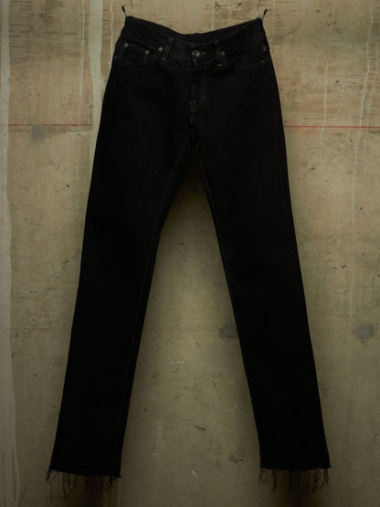 Black denim jeans with a raw, frayed hem, hanging against a textured concrete wall.