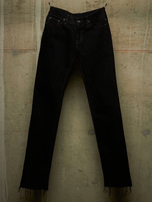 Black denim jeans with a raw, frayed hem, hanging against a textured concrete wall.