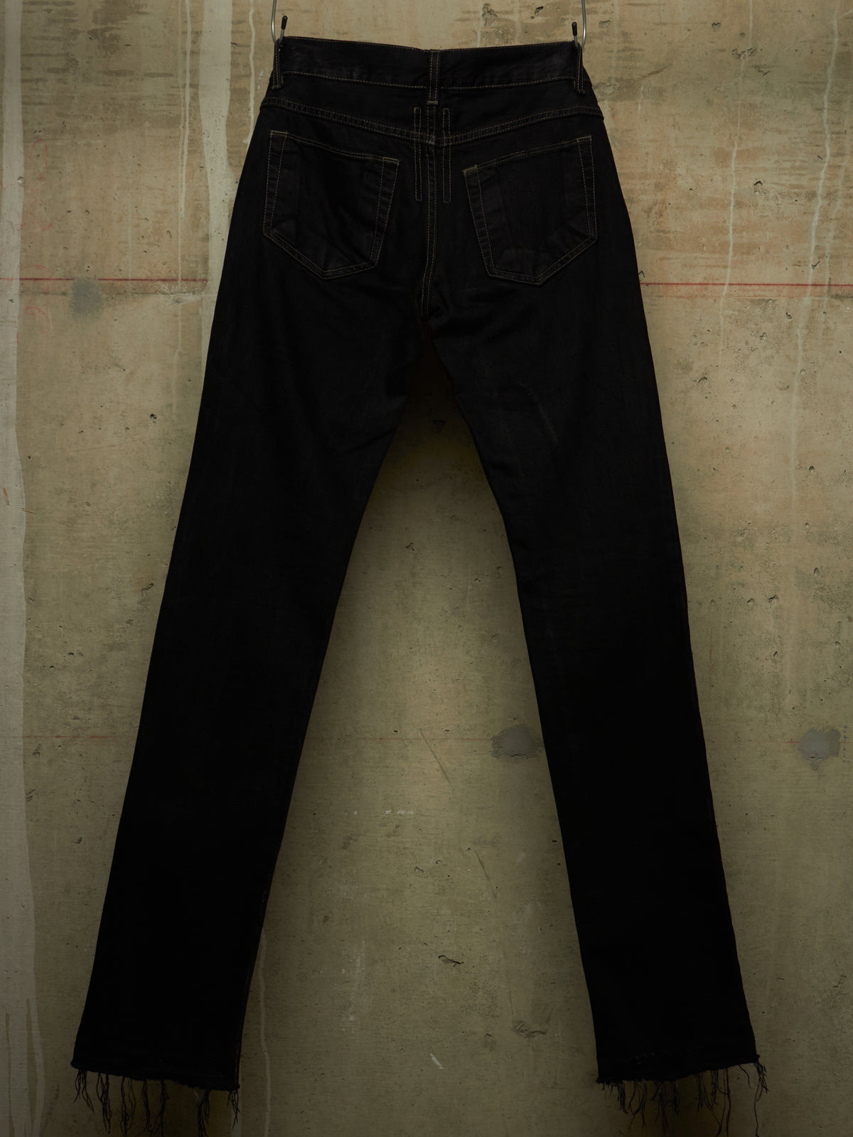 Black denim jeans shown from the back, hanging against a textured concrete wall. The back pockets are visible, and the hems have a frayed, raw edge.