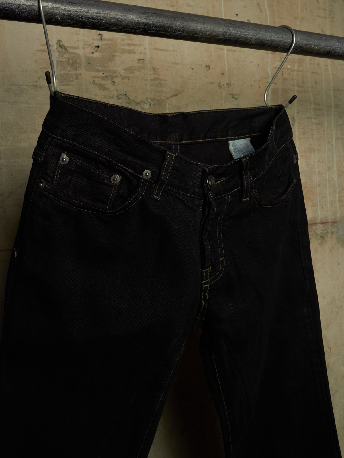 Black denim jeans hanging from a metal hanger on a dark metal rod against a textured beige wall.