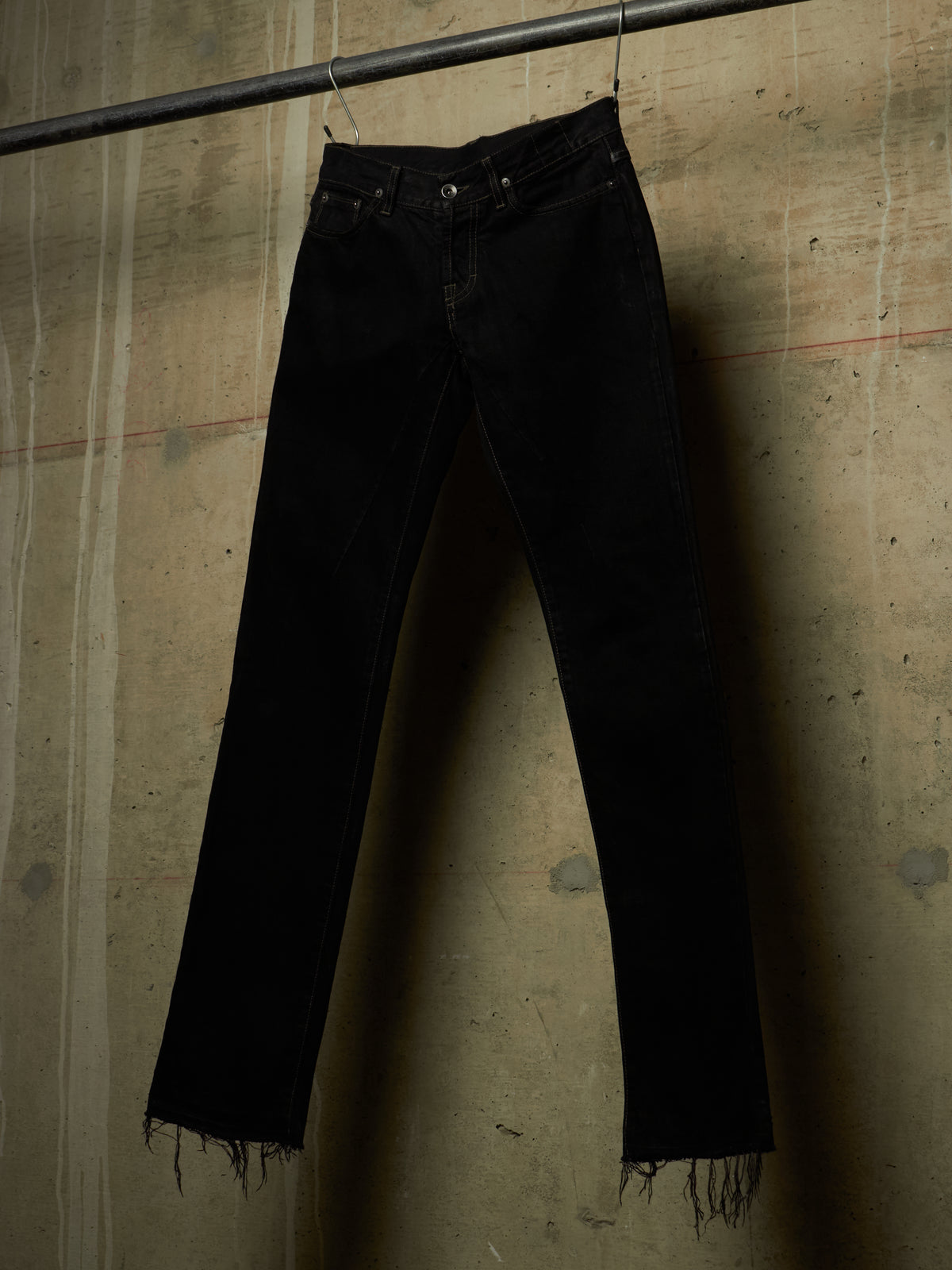 Black denim jeans with raw, frayed hems hanging from a metal rod against a textured concrete wall.