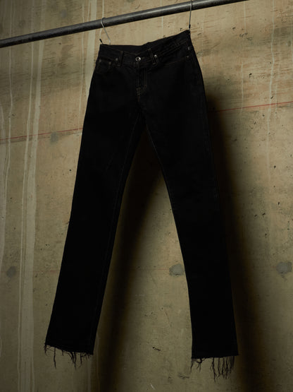 Black denim jeans with raw, frayed hems hanging from a metal rod against a textured concrete wall.