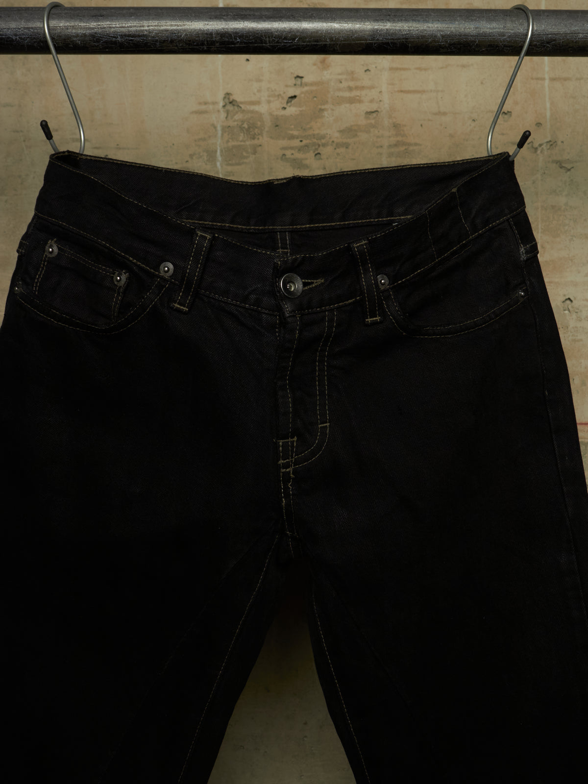 Black denim jeans hanging from a metal hanger on a dark rod against a textured beige wall.