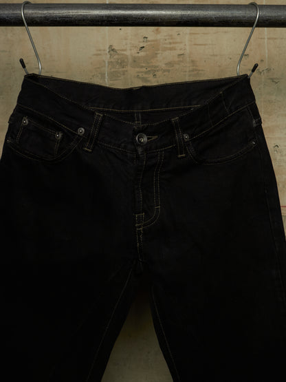 Black denim jeans hanging from a metal hanger on a dark rod against a textured beige wall.