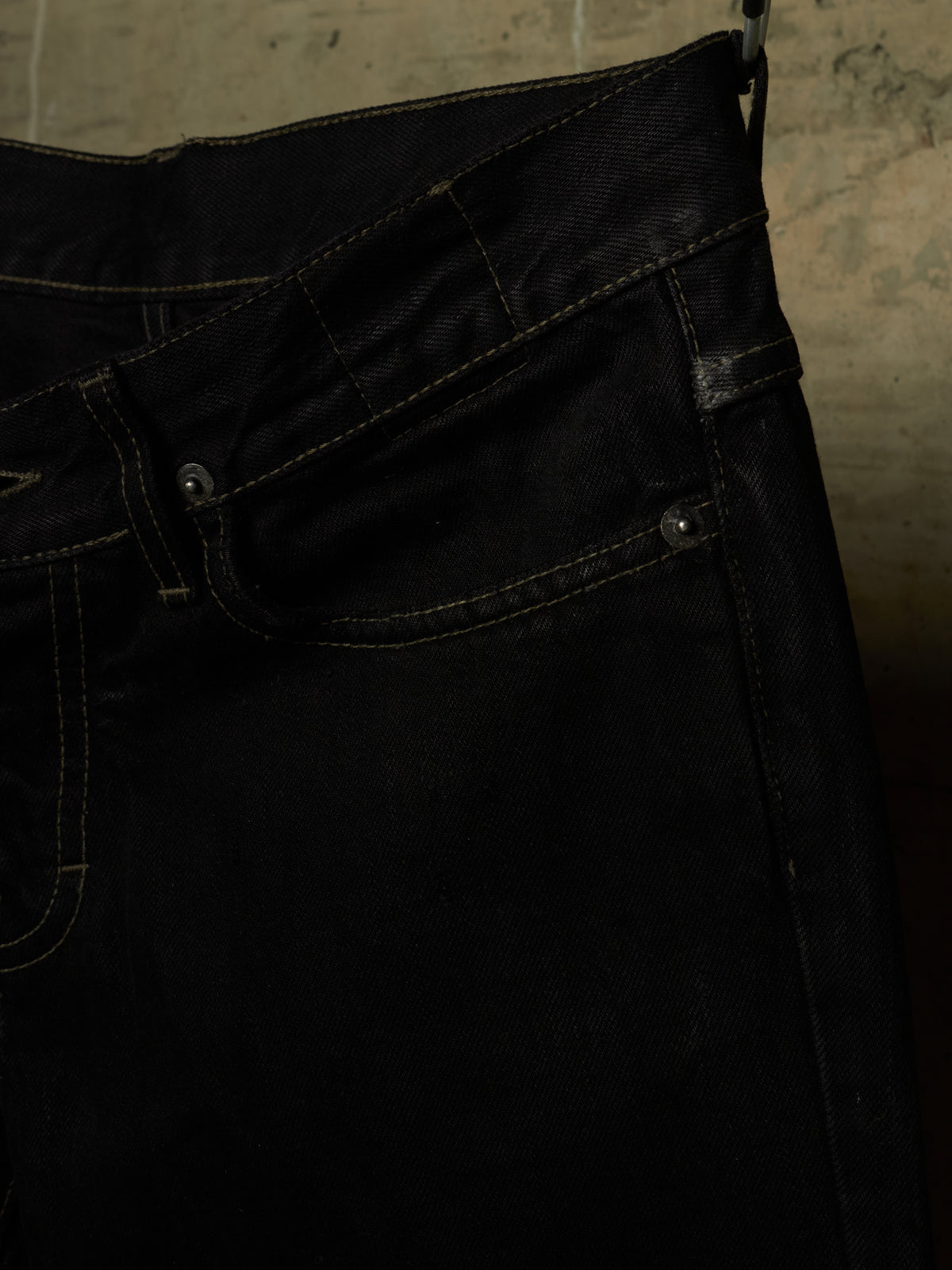 Close-up of a pair of dark indigo or black denim jeans showing the waistband, belt loops, front pocket, and rivets against a textured concrete background.