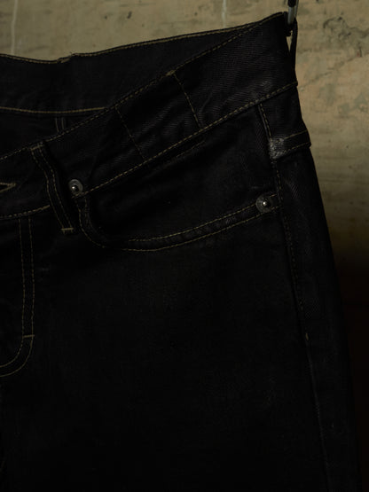 Close-up of a pair of dark indigo or black denim jeans showing the waistband, belt loops, front pocket, and rivets against a textured concrete background.