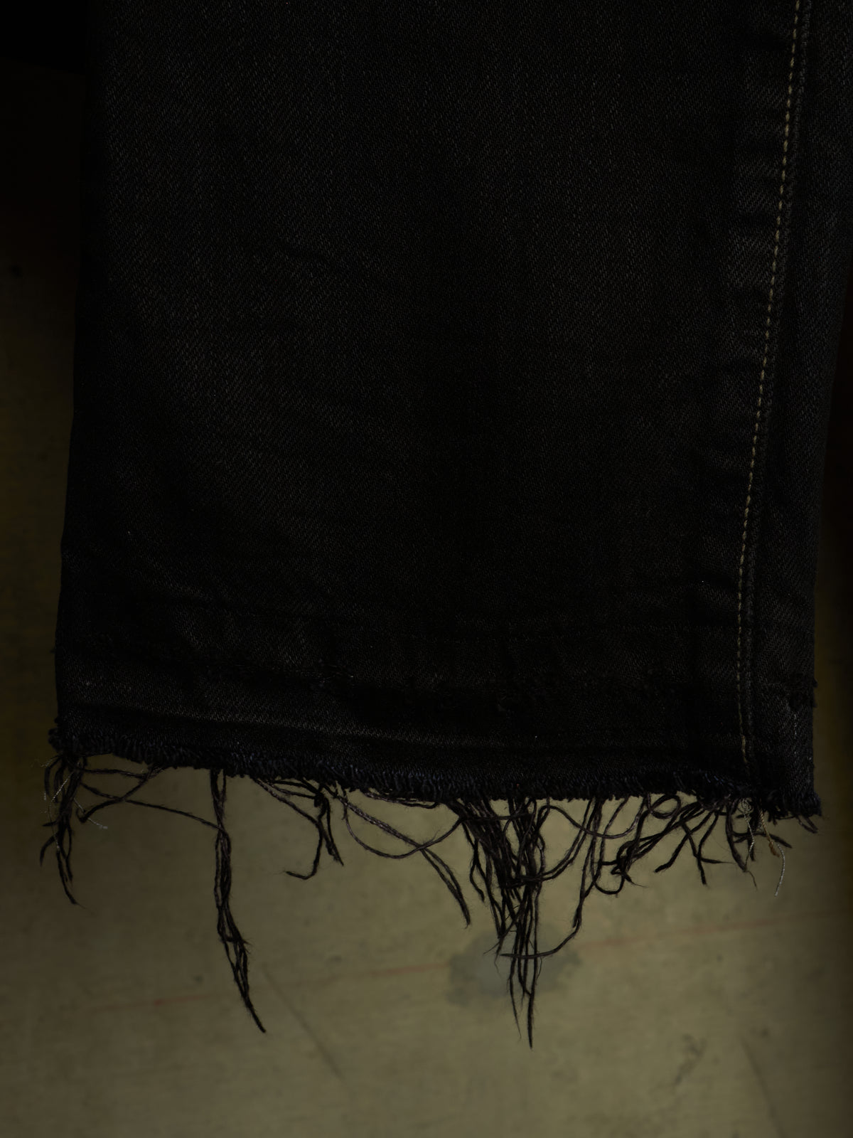 Close-up of a black frayed denim fabric, showing the raw, frayed edge and subtle contrast stitching against a dark background.