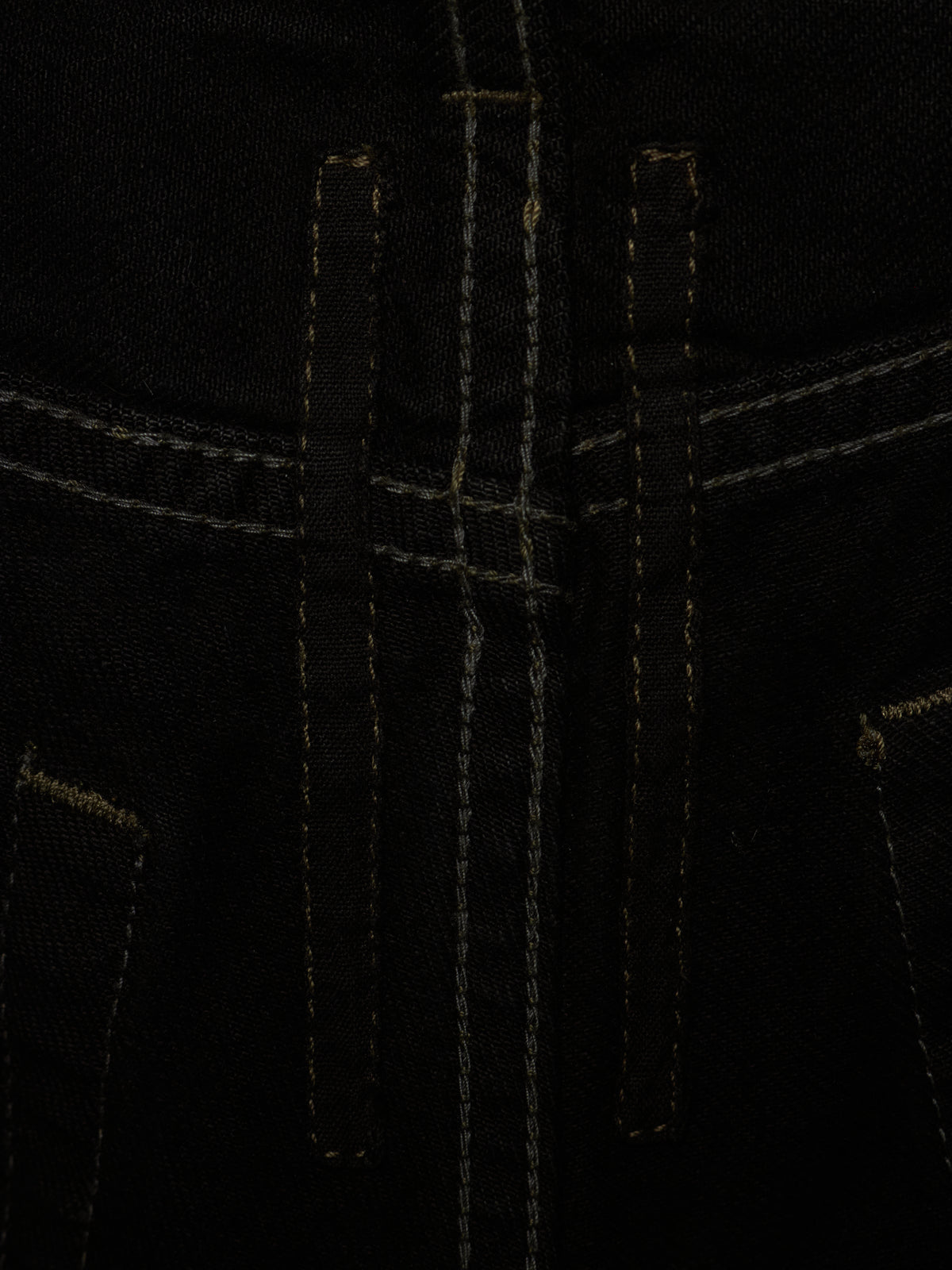 Close-up detail of black denim fabric with contrast light tan stitching, showing the front fly area and pocket edges.