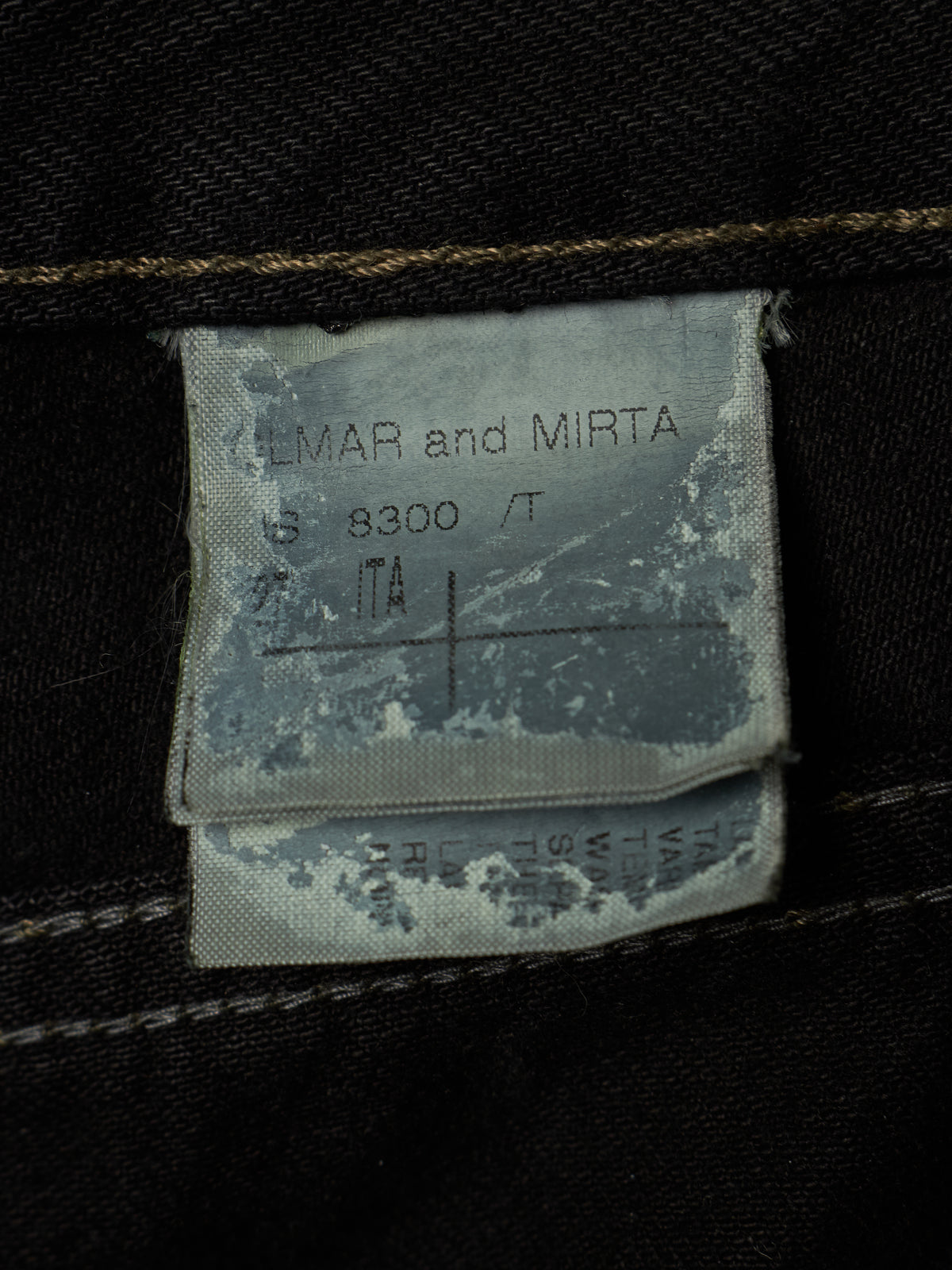 Close-up of a faded gray fabric garment label sewn onto dark black denim, showing the text 'LMAR and MIRT A 8300 / T' and size information.
