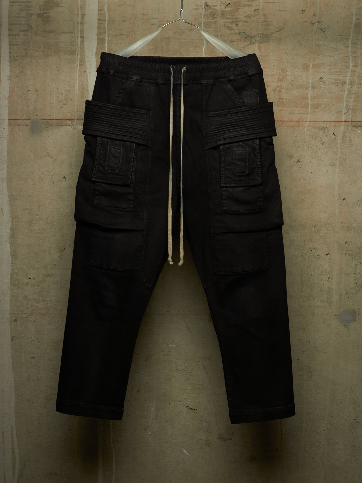 Black cargo pants with a drawstring waist and multiple flap pockets, hanging against a textured concrete wall.