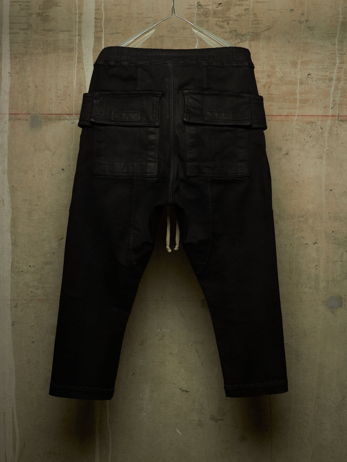 Black cargo-style pants with a drawstring waist and flap cargo pockets, hanging against a textured concrete wall.