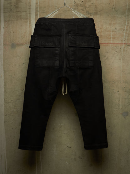 Black cargo-style pants with a drawstring waist and flap cargo pockets, hanging against a textured concrete wall.