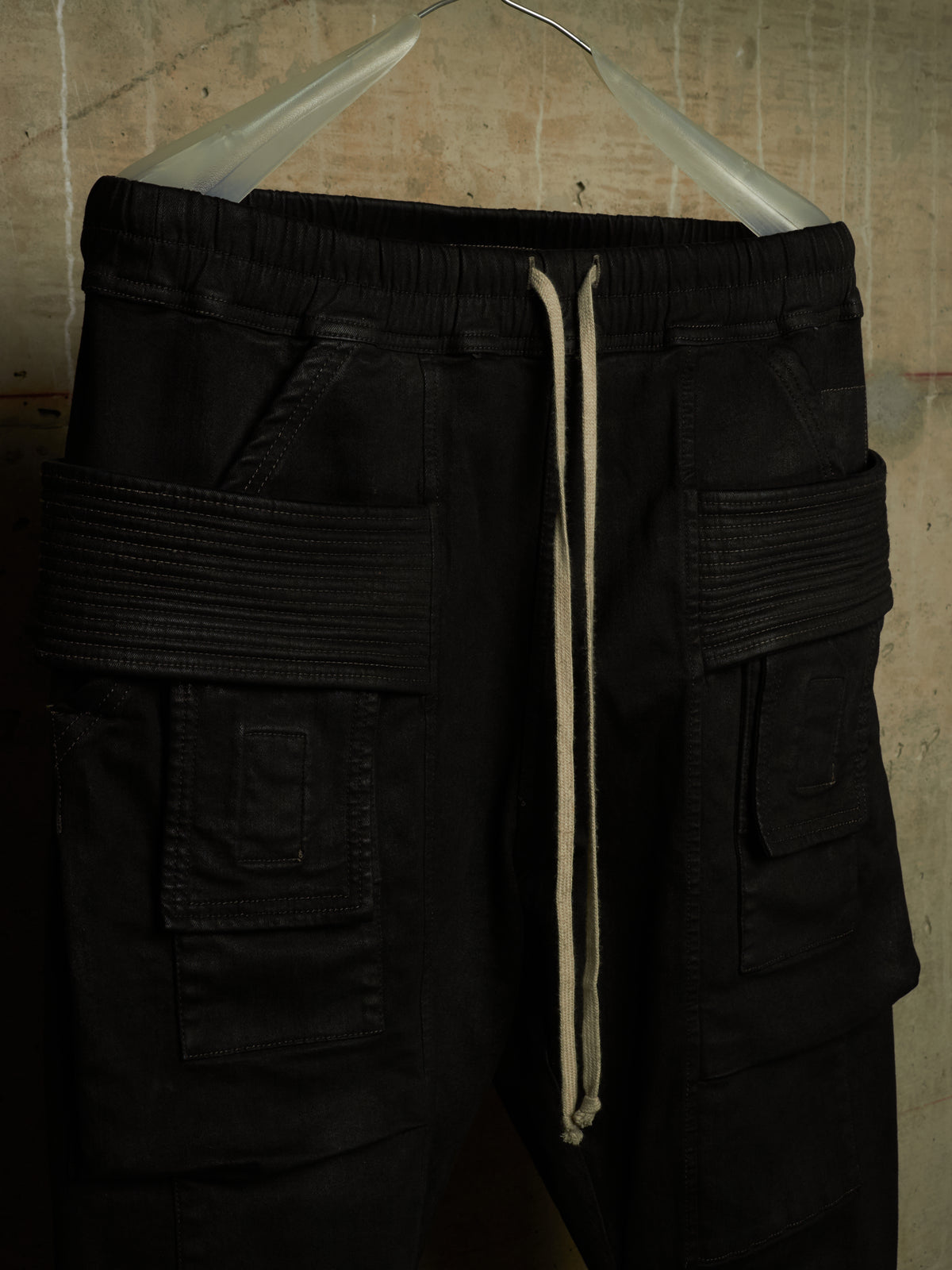 Black cargo-style pants with a cream drawstring, hanging on a clear plastic hanger against a textured concrete wall.