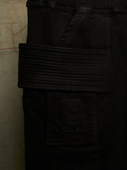 Close-up detail of black cargo pants showing a wide, ribbed elastic belt-like strap across the pocket and a stitched flap pocket below, photographed against a textured neutral background.