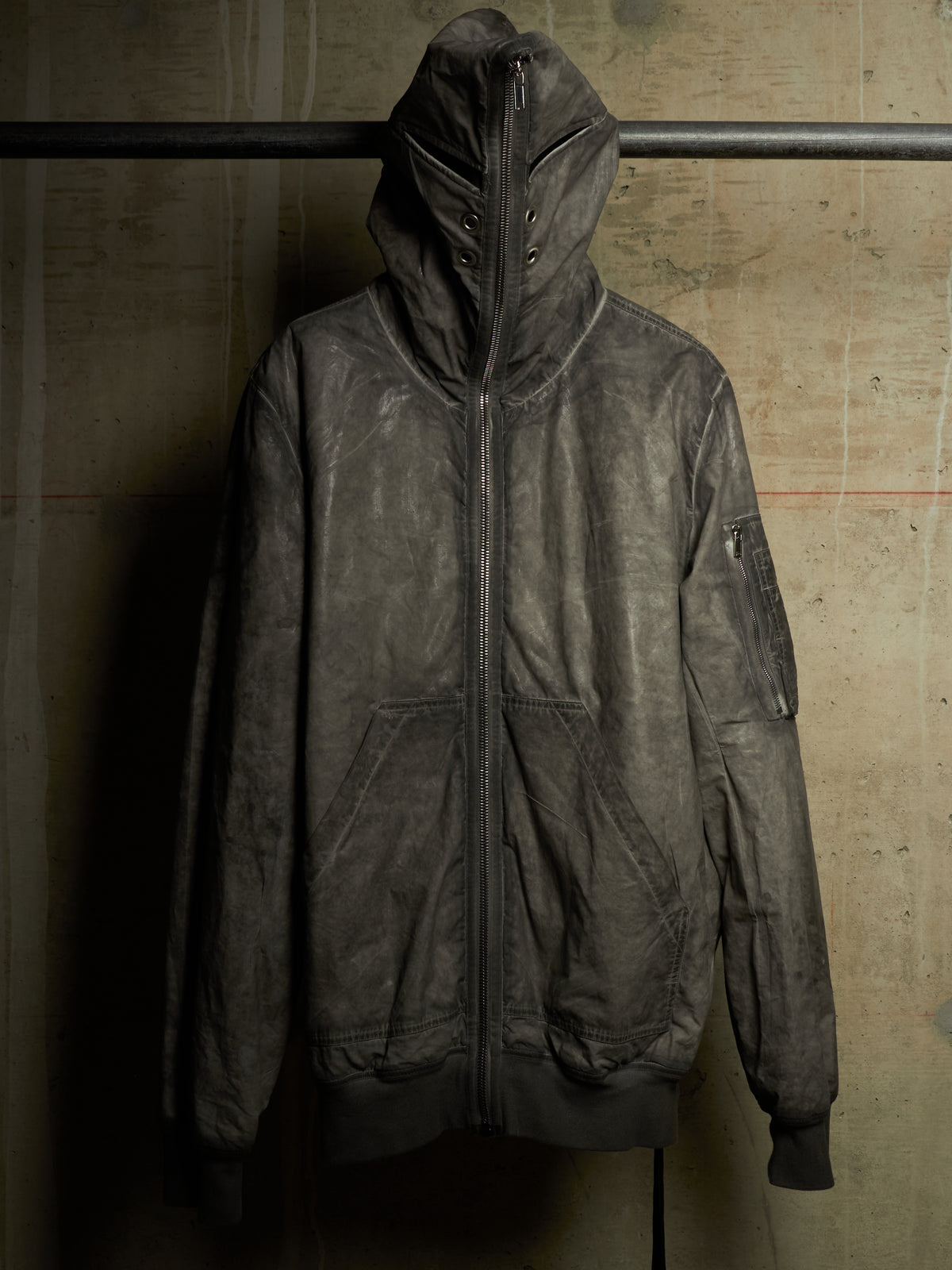 A distressed, dark gray leather hooded jacket with a front zipper, hanging on a black metal rack against a textured concrete wall.