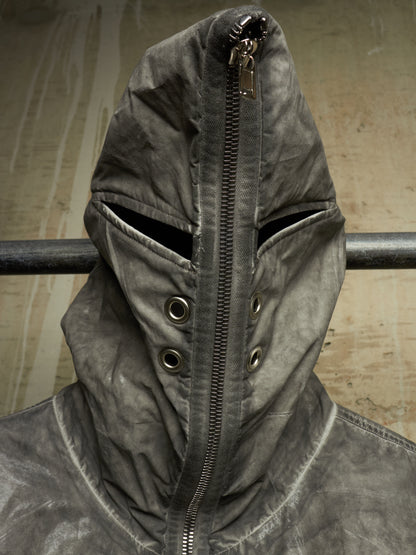 A close-up of a dark gray hooded jacket with a front zipper, eye slits, and metal snap details, shown against a textured beige background.