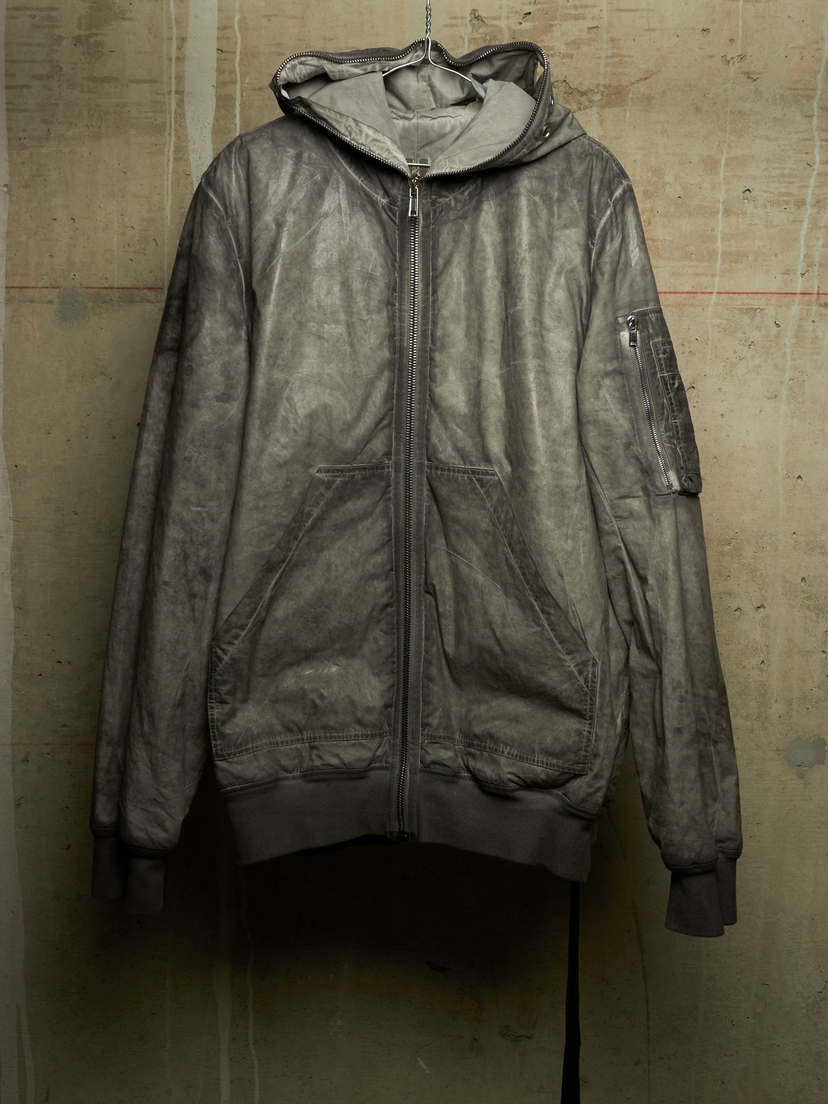 A distressed, washed-out dark gray hooded zip-up jacket with front pockets, hanging against a textured concrete wall.