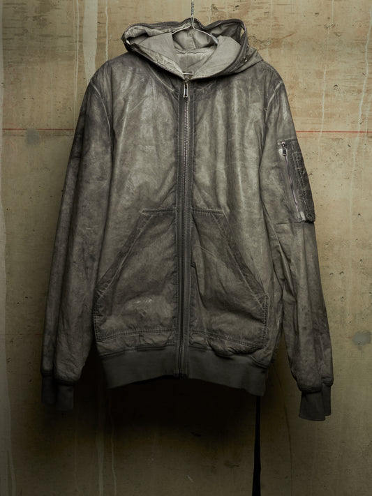 A distressed, washed-out dark gray hooded zip-up jacket with front pockets, hanging against a textured concrete wall.