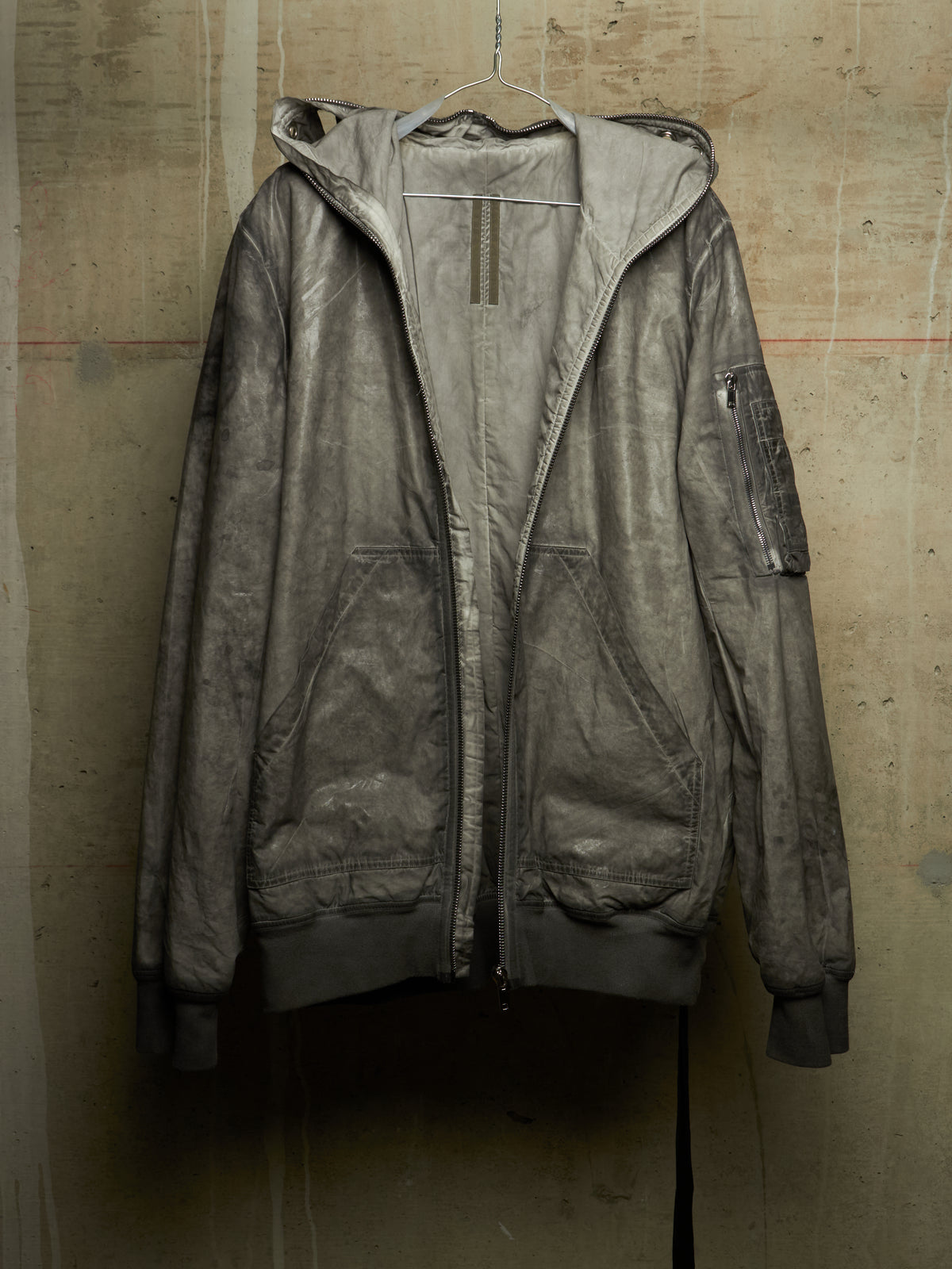 A distressed, washed-out gray hooded jacket hanging on a wire hanger against a textured concrete wall.
