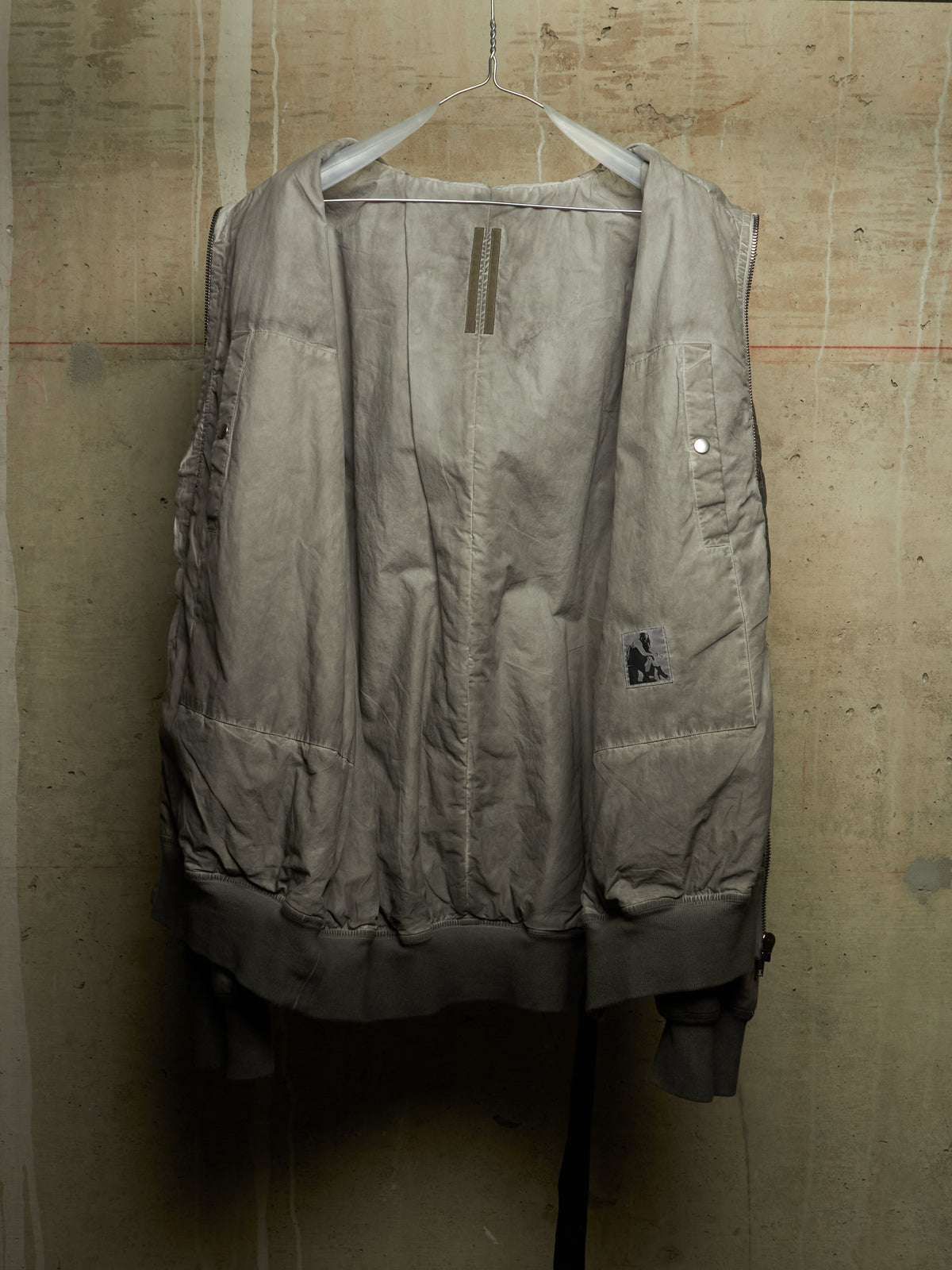 A light gray quilted puffer vest hanging on a wire hanger against a textured concrete wall. The vest features a front zipper, a small patch on the left chest pocket, and a small embroidered logo on the right pocket.