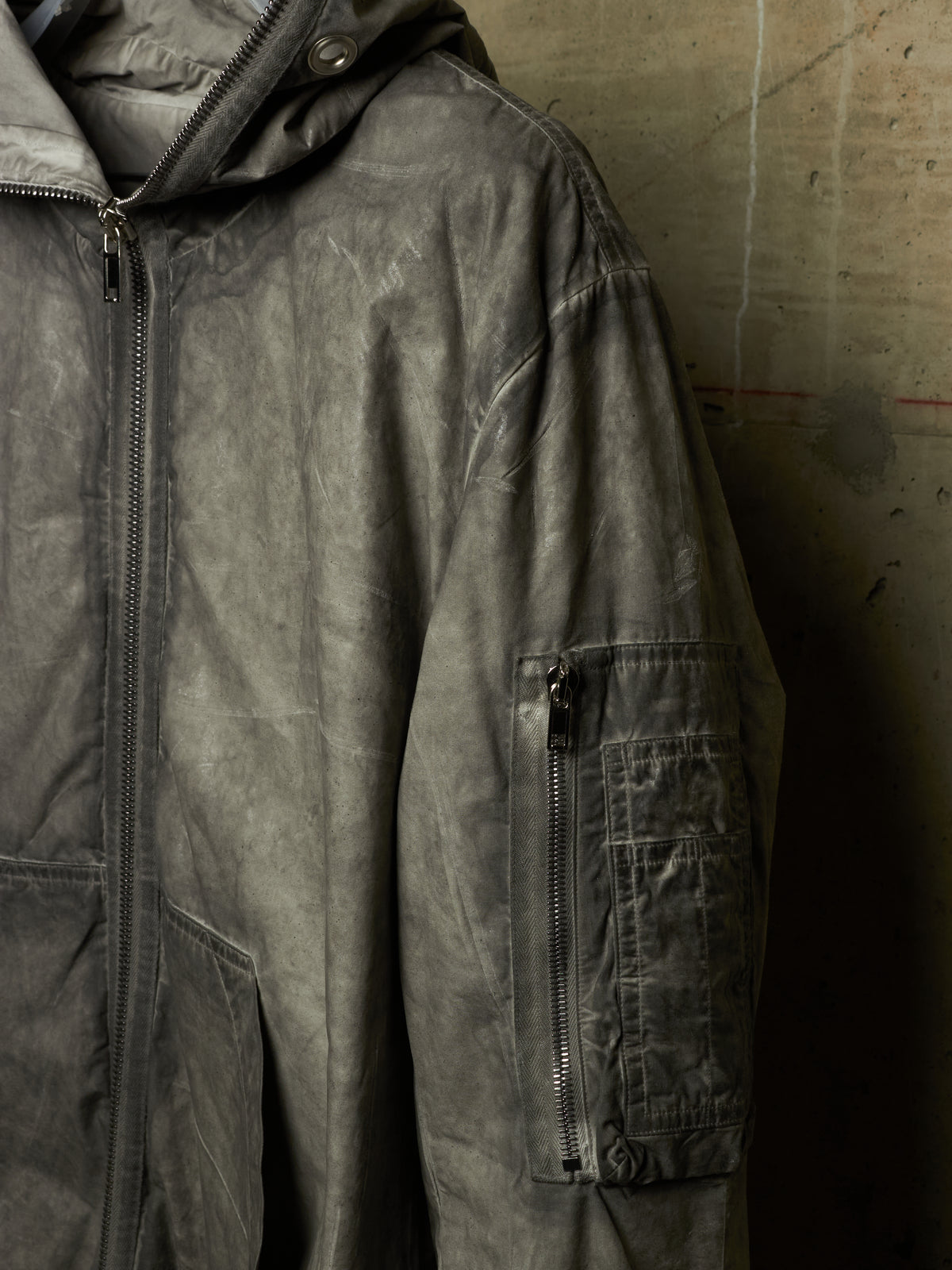 A close-up of a dark gray hooded jacket with a distressed, washed texture, featuring a vertical zippered pocket on the sleeve, set against a concrete wall.