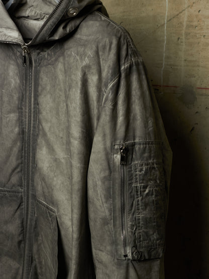 A close-up of a dark gray hooded jacket with a distressed, washed texture, featuring a vertical zippered pocket on the sleeve, set against a concrete wall.