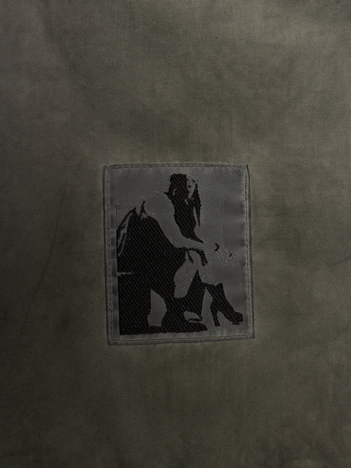 A close-up of a dark olive green textured fabric featuring a rectangular embroidered patch with a black silhouette graphic of a person sitting on a horse.