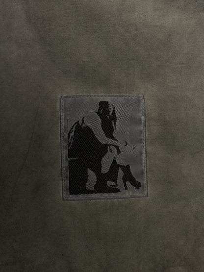 A close-up of a dark olive green textured fabric featuring a rectangular embroidered patch with a black silhouette graphic of a person sitting on a horse.