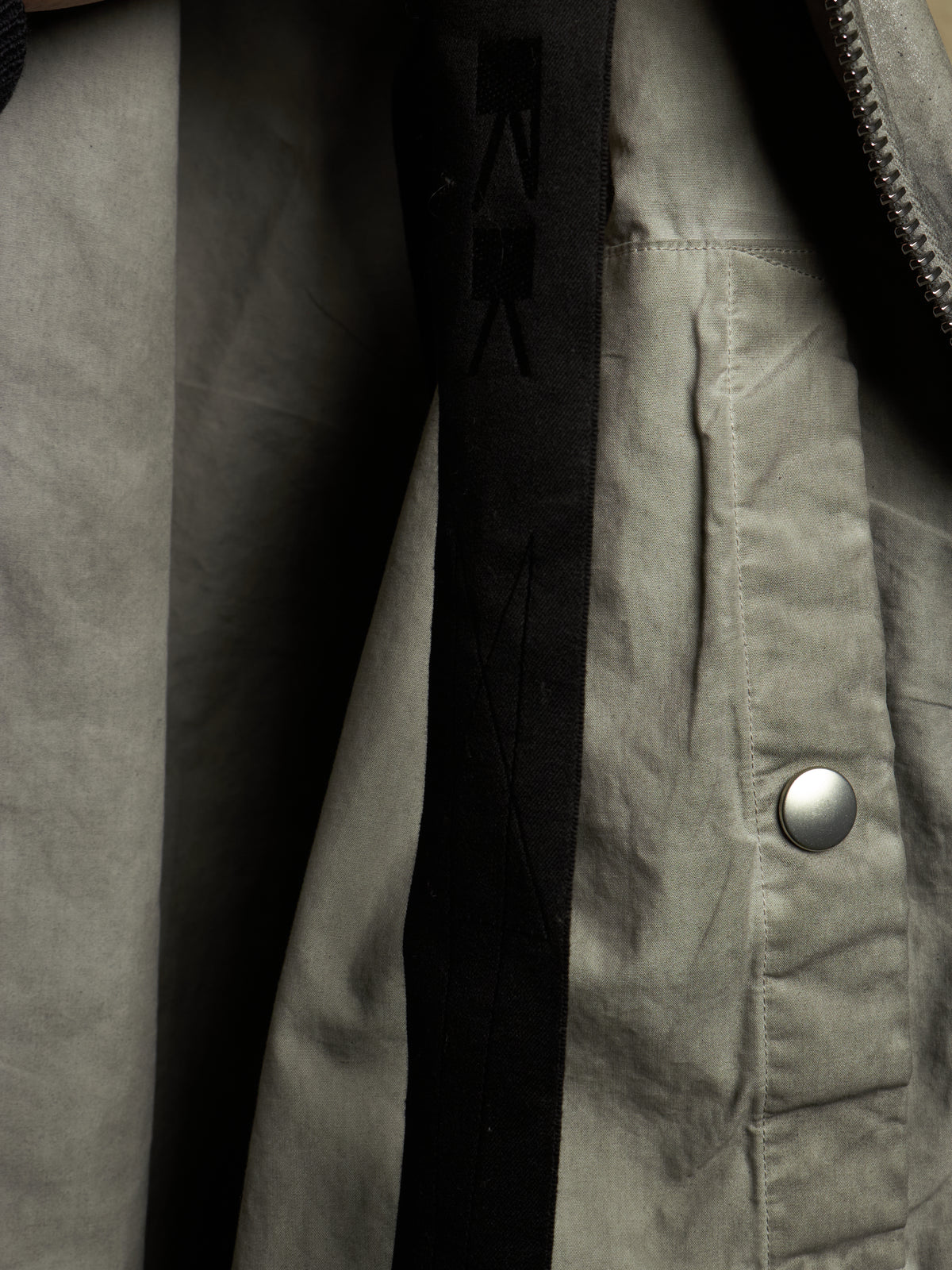 Close-up detail of a jacket showing a light gray fabric exterior, a black vertical inner panel with tonal logo stitching, a silver snap button, and a zipper.